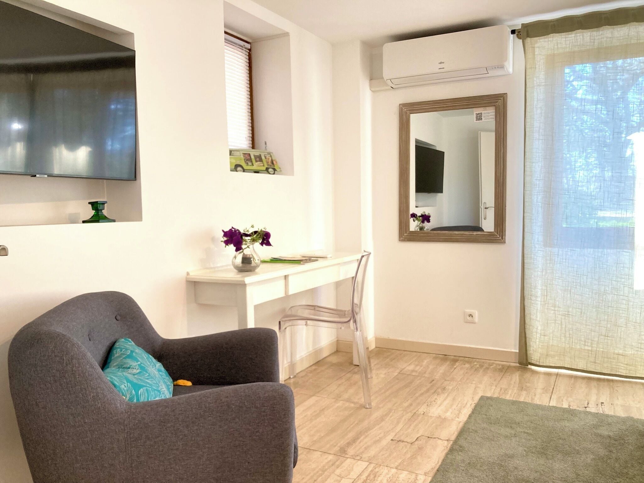A living room with a grey sofa, a white desk, a clear chair, a flat-screen TV mounted on the wall, and a large mirror.