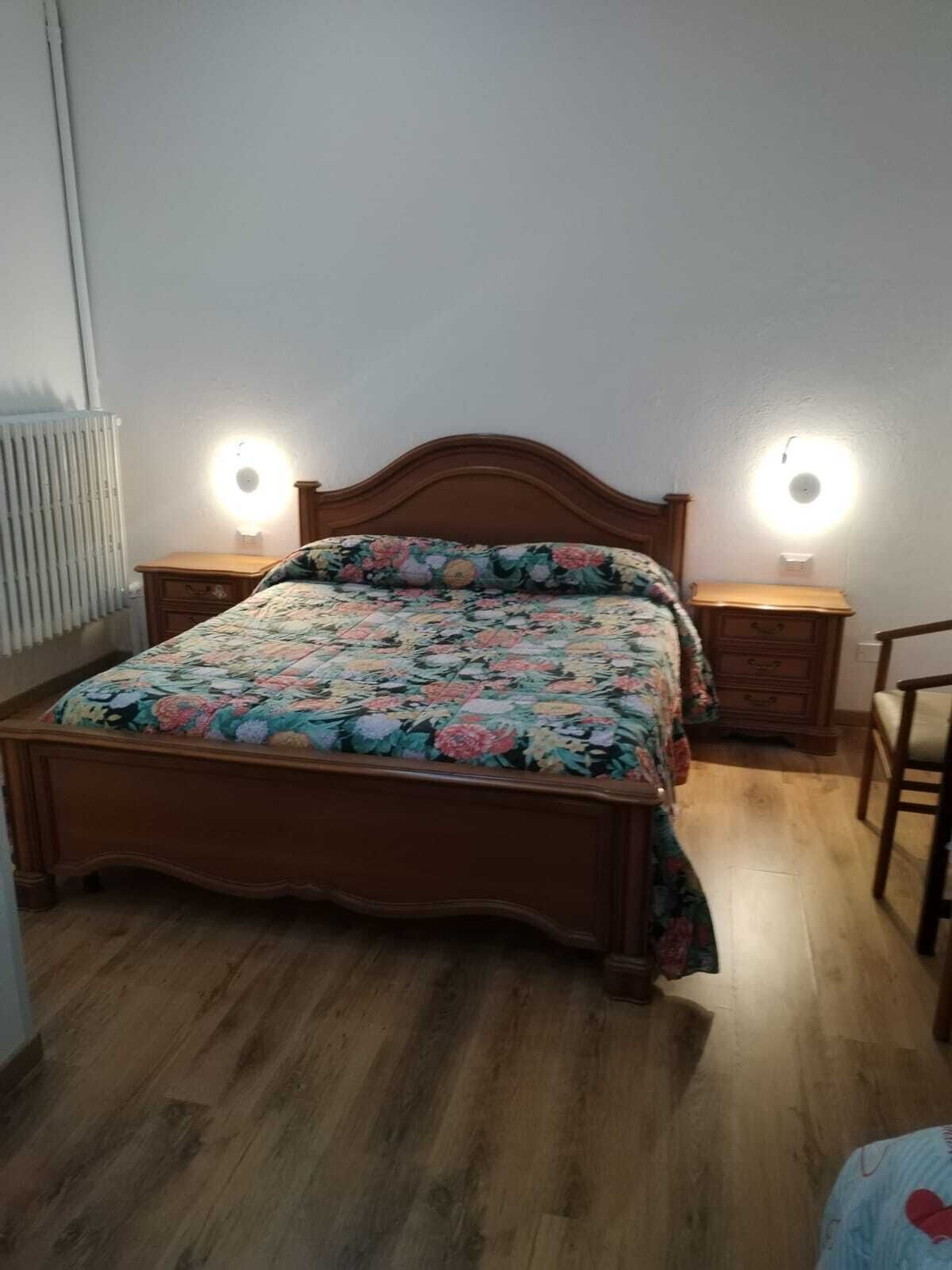 A bedroom with a wooden bed, floral bedding, two bedside tables with lamps, and a radiator.
