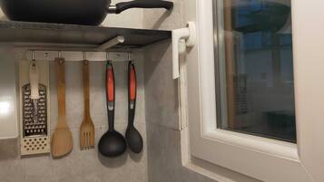 A kitchen with a stove, a pot on the stove, a hanging pot rack with various utensils, and a window with a view of the outside.