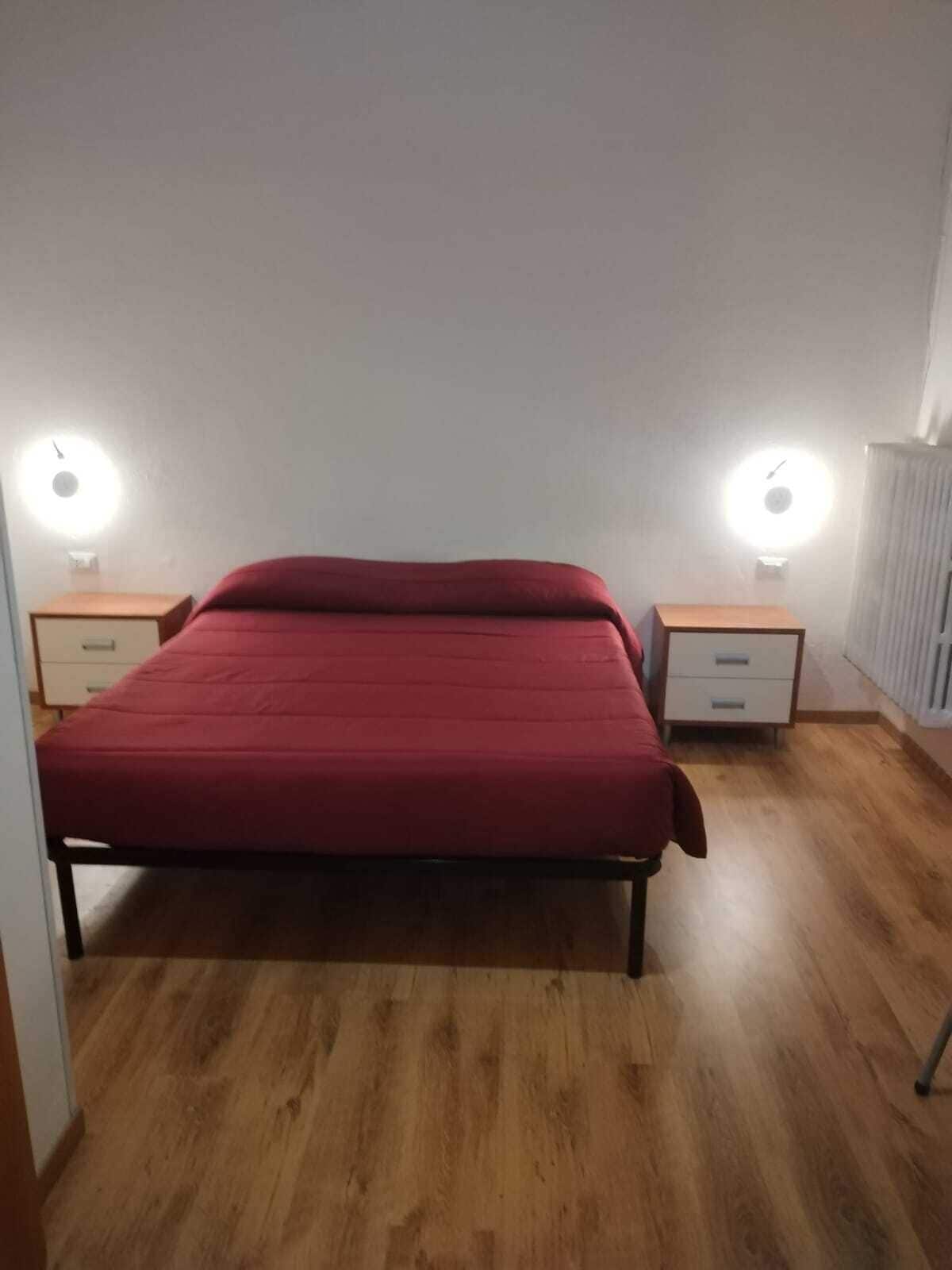 A single-bed room with a wooden floor, a red bedspread, two bedside tables with lamps, and a white radiator.
