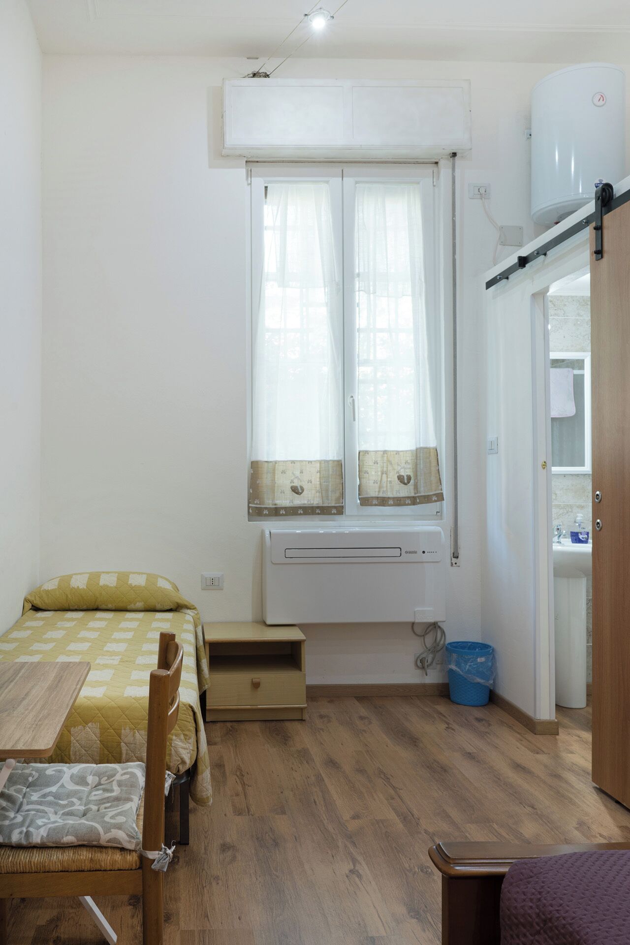 A small, well-lit room with a bed, a table, and a window with curtains.