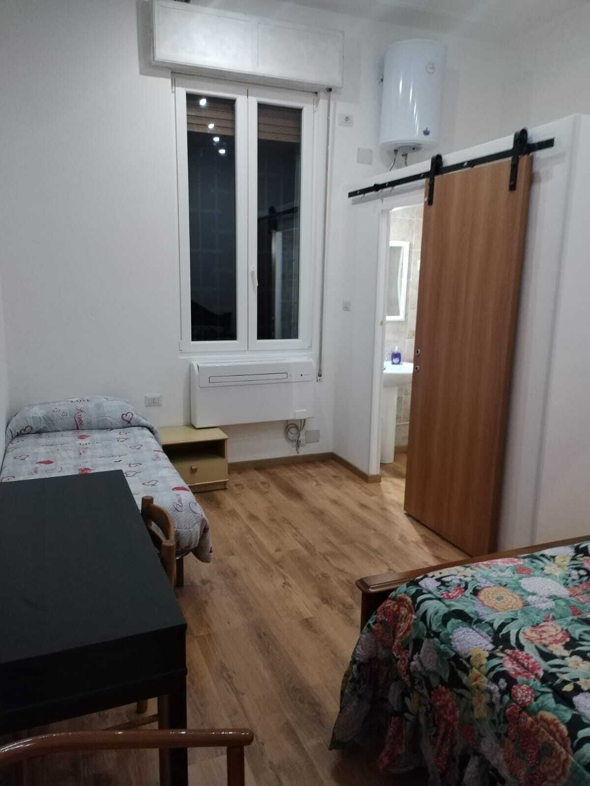 Ein kleiner, gut beleuchteter Raum mit einem Bett, einem Tisch und einem Stuhl. Es gibt ein Fenster mit Blick hinaus, eine Tür zu einem weiteren Zimmer und eine Heizung an der Wand.