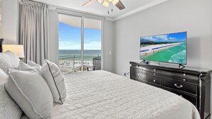 Une chambre avec un grand lit, un ventilateur de plafond, un téléviseur posé sur une commode et une vue sur la plage, visible à travers une porte-fenêtre coulissante.