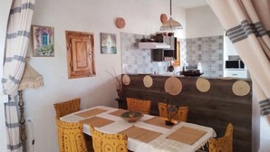 A dining area with a table set for four, surrounded by patterned chairs, a kitchen in the background, and a hanging light fixture. - Chambre D'hôte 2personnes Avec Petit Déjeuner (Mednine)