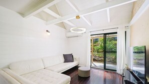 Un salon moderne avec un canapé blanc, un pouf rond, un luminaire suspendu au plafond et une porte-fenêtre coulissante donnant sur un jardin.