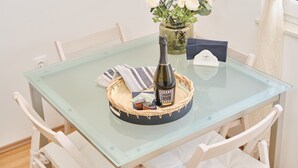 A glass-top table with a bottle of champagne, a vase with flowers, and a card on a woven coaster.