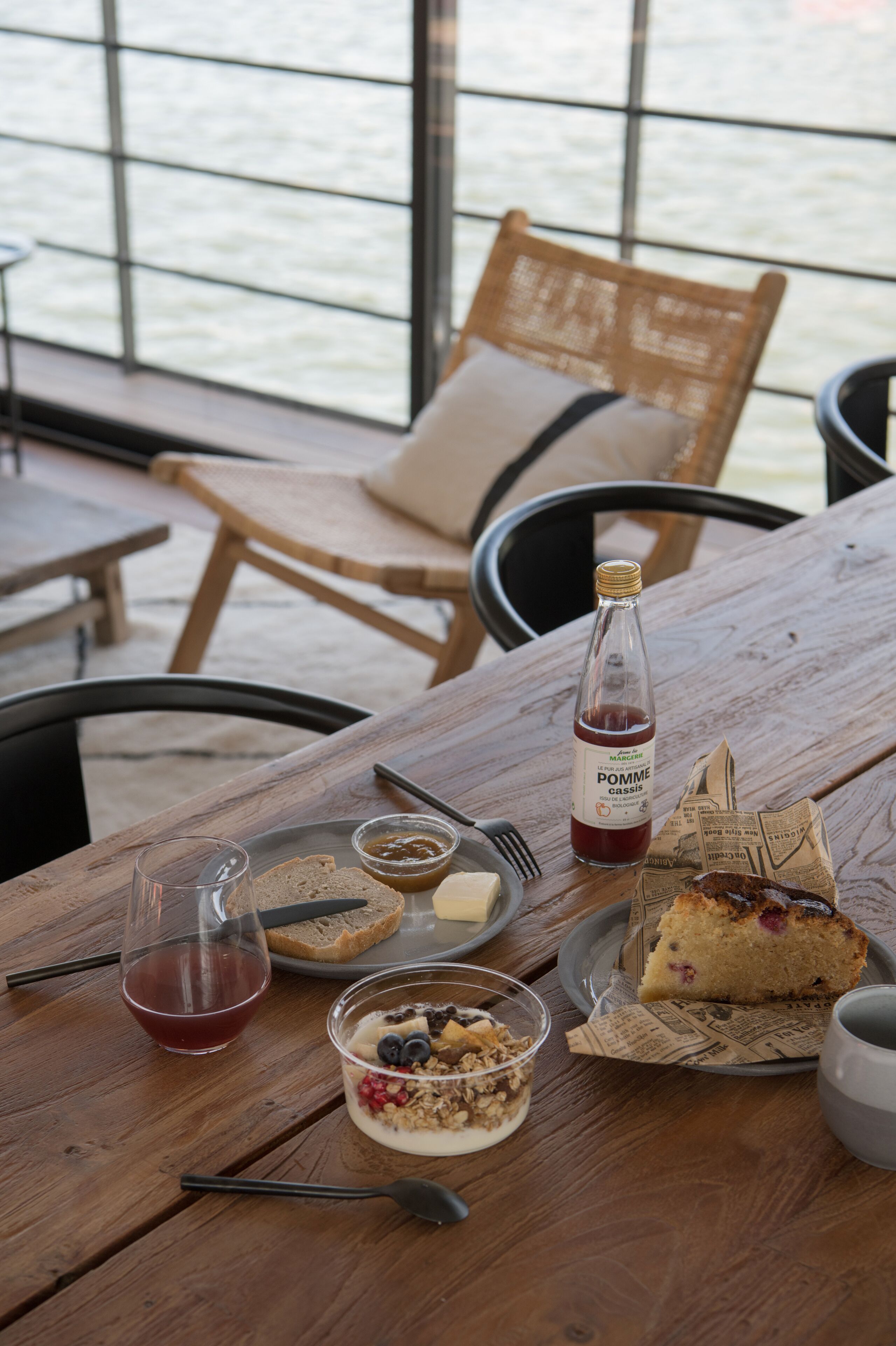 A wooden table with a meal consisting of bread, jam, a bottle of Pomme cassis, a glass of tea, a bowl of yogurt with berries, and a slice of cake.
