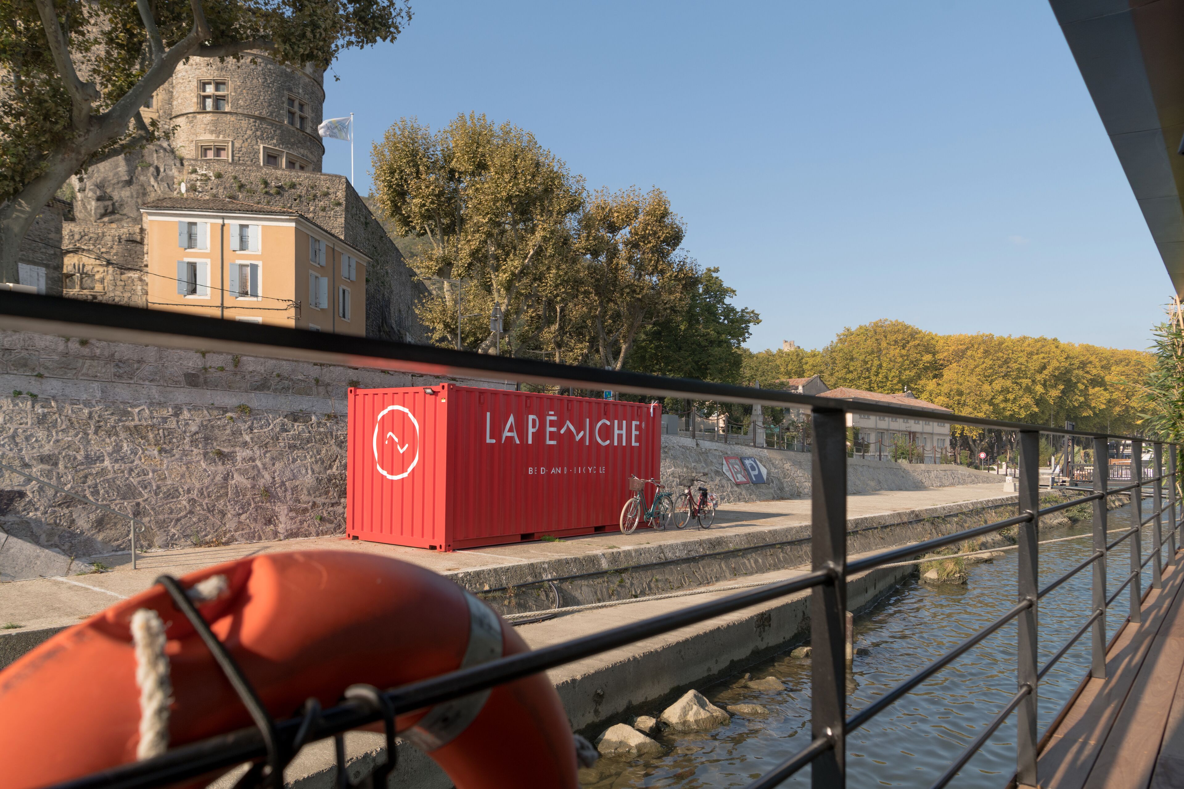 A red shipping container labeled "LAPEMCHE" on a dock with bicycles and a stone wall in the background.