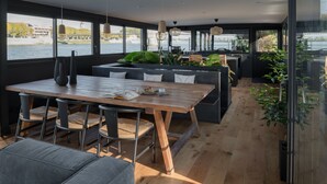 A modern interior with a wooden dining table, black upholstered chairs, and a sofa. There are large windows offering a view of the water and a bridge. - LA PENICHE - Bed And Bicycle - Tournon (Tournon-sur-Rhone)