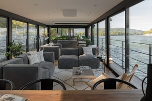 Un espacio habitable moderno y de planta abierta con un amplio ventanal que da a una masa de agua, amoblado con sofás y una mesa de centro.
