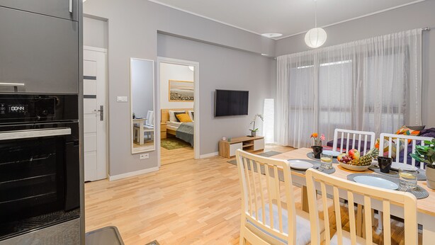 Une cuisine moderne dotée d’un four encastré, un coin repas avec une table et des chaises en bois, et un salon avec un lit et un téléviseur.