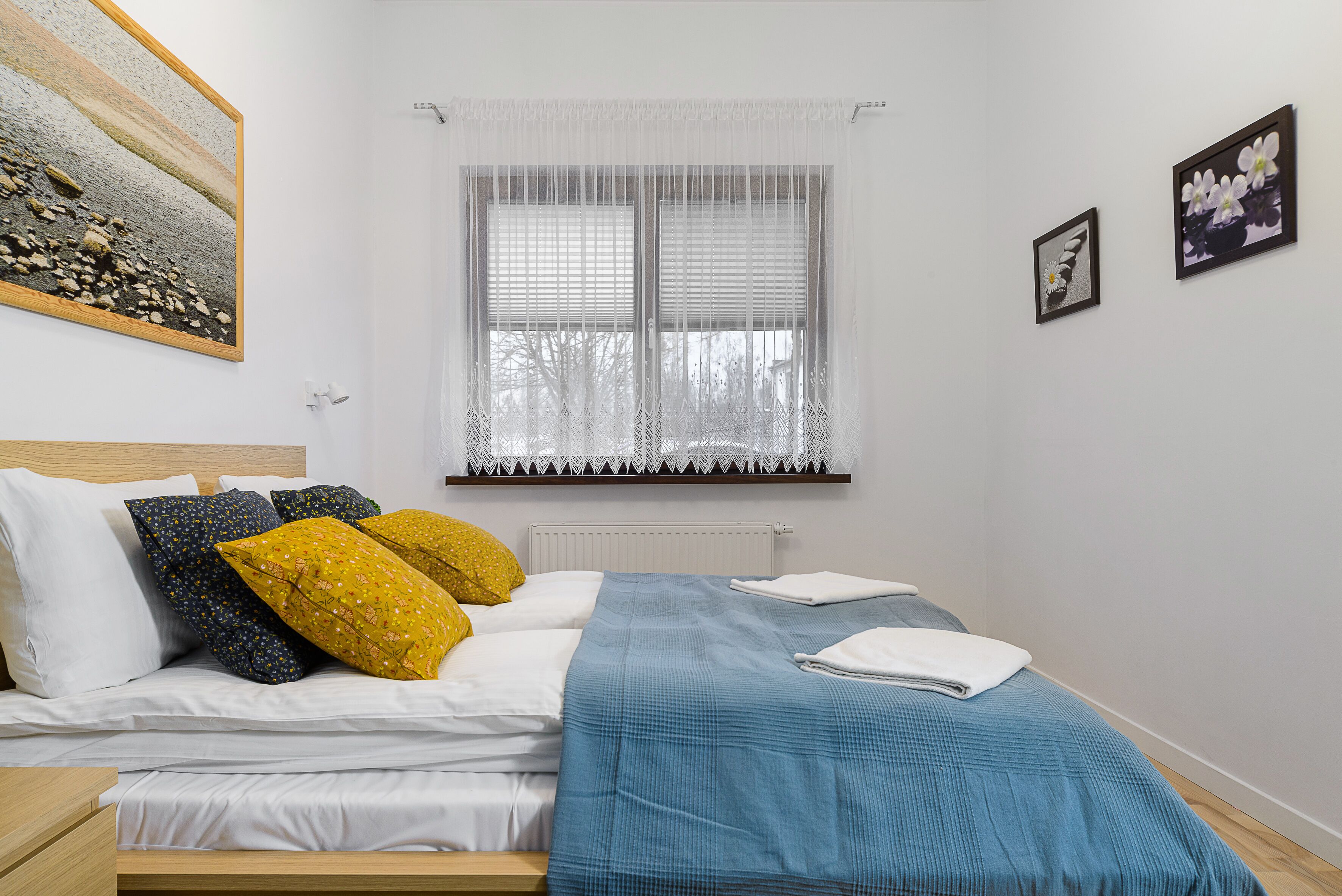 A neatly made bed with white and blue bedding, a window with sheer curtains, and two framed pictures on the wall.