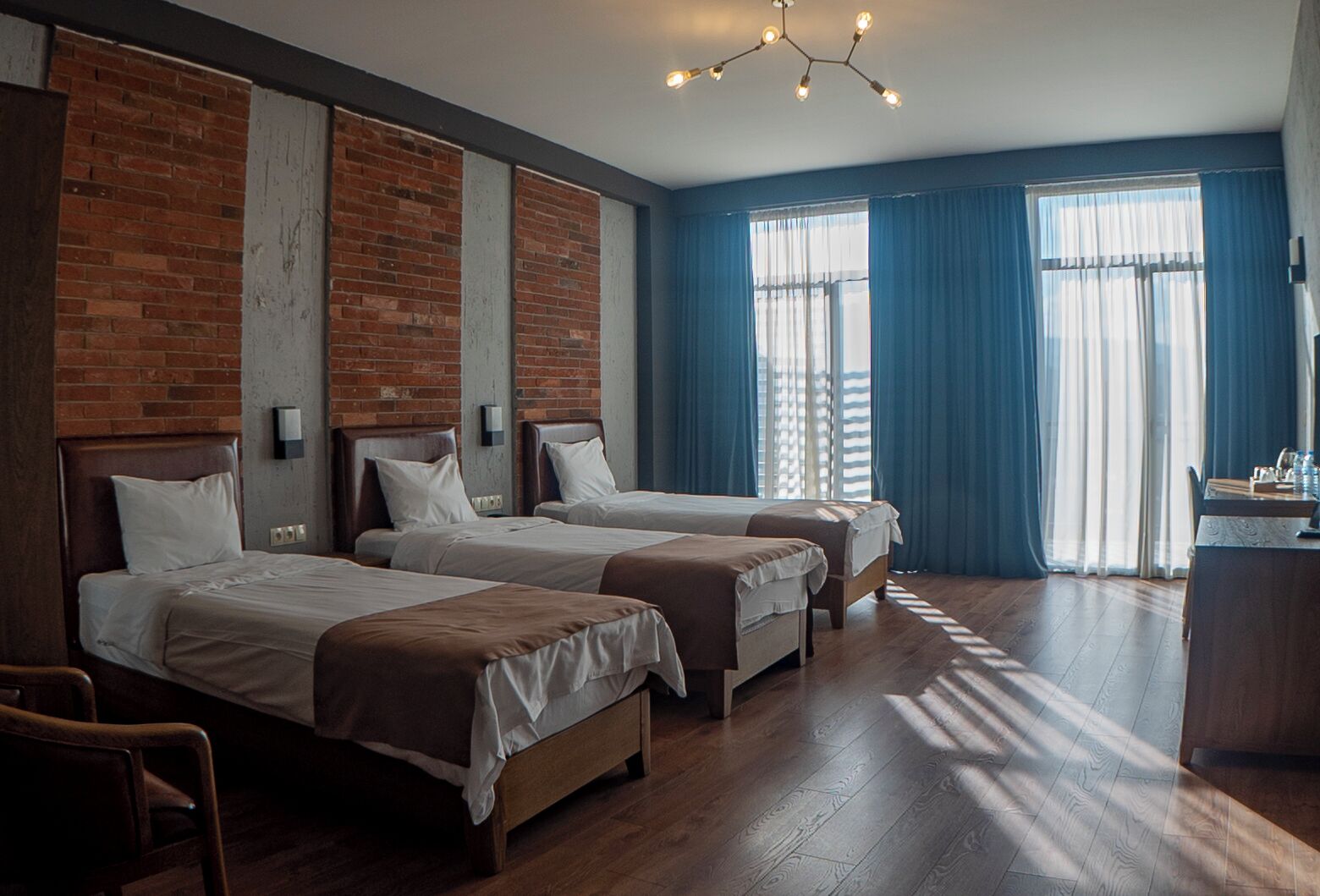 A hotel room with three single beds, a wooden floor, a brick accent wall, and a desk with a chair.