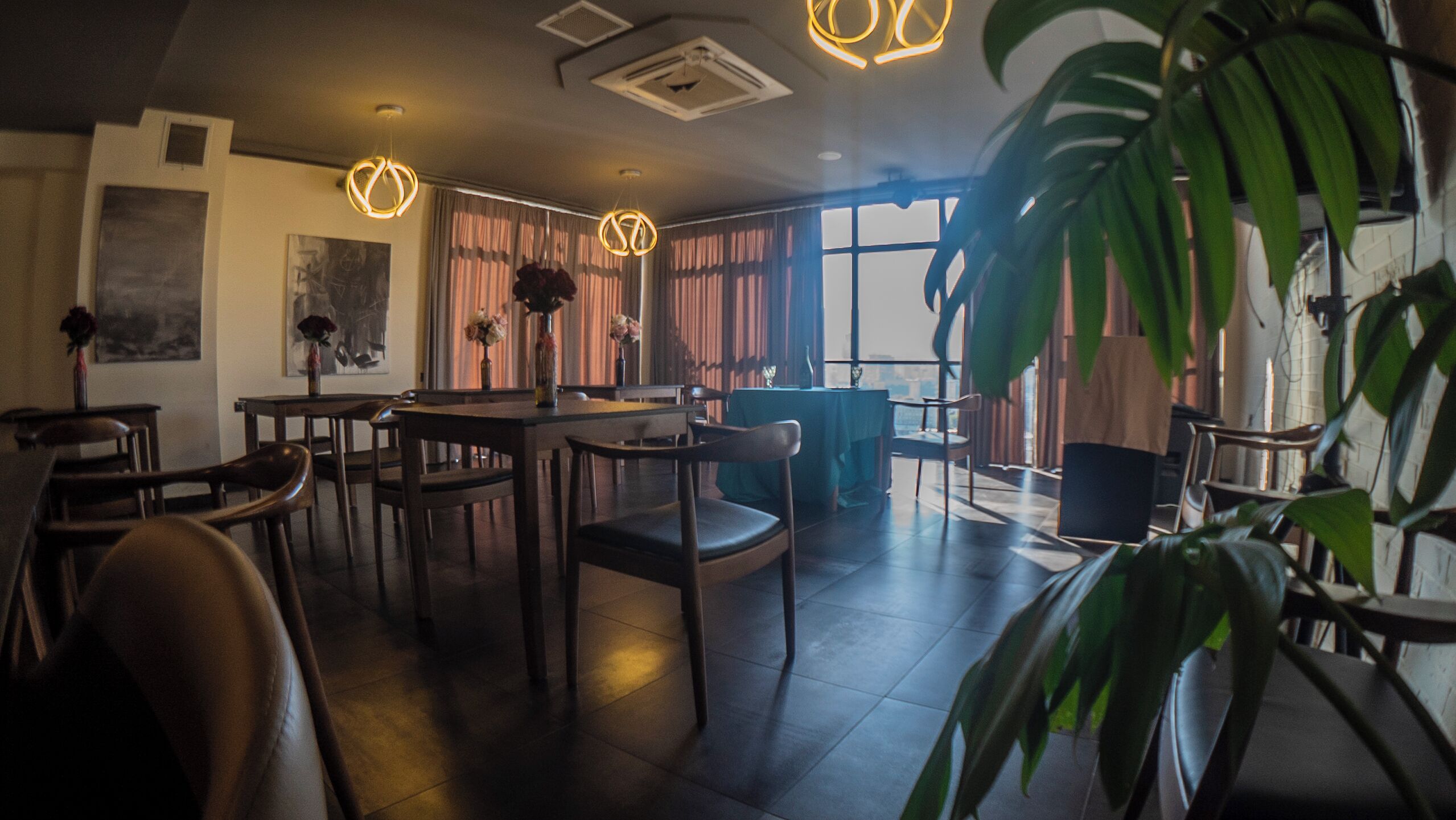 A modern dining area with wooden tables and chairs, pendant lighting, and large windows with curtains.
