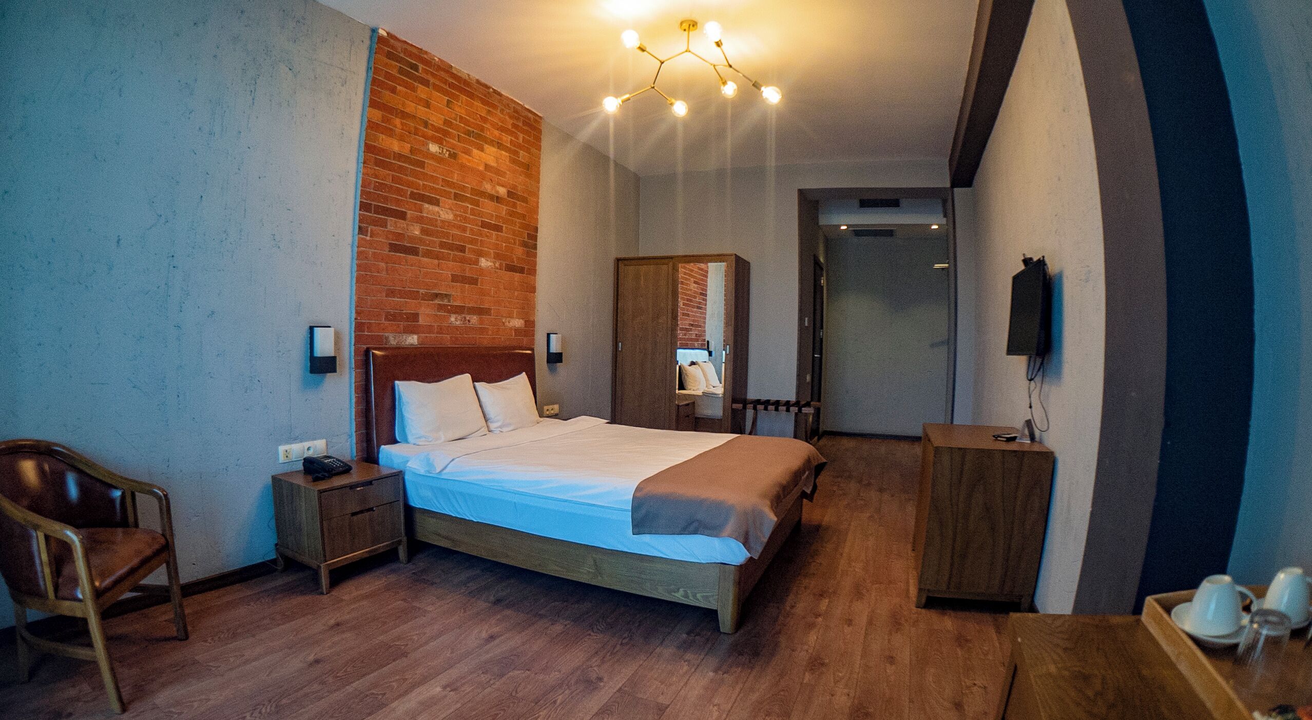 A hotel room with a wooden floor, a bed with white linens, a wooden nightstand, a chair, a wooden wardrobe, and a television on the wall.