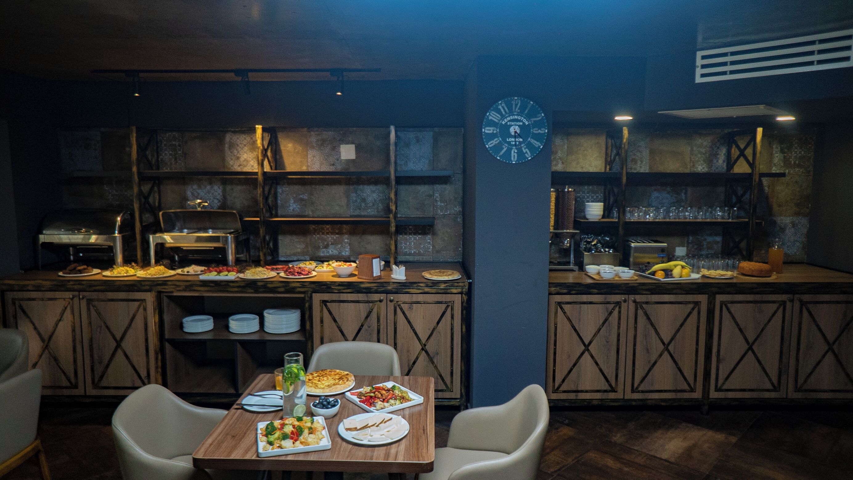 Un ristorante con un'atmosfera rustica, caratterizzato da mobili in legno, una sveglia appesa al muro e diverse portate esposte sul bancone.