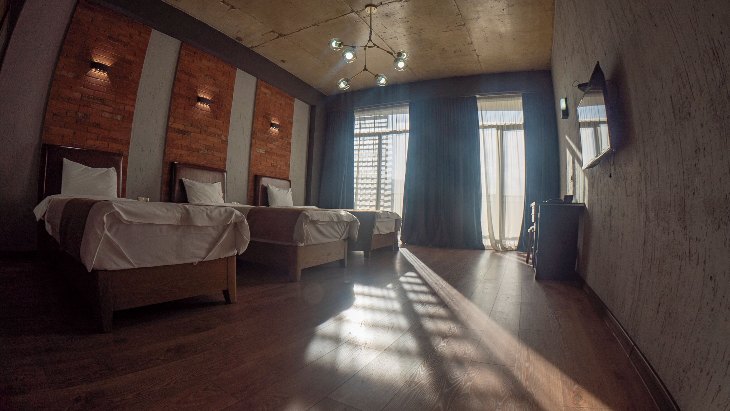 A hotel room with two beds, a brick wall, wooden flooring, and a large window with curtains.