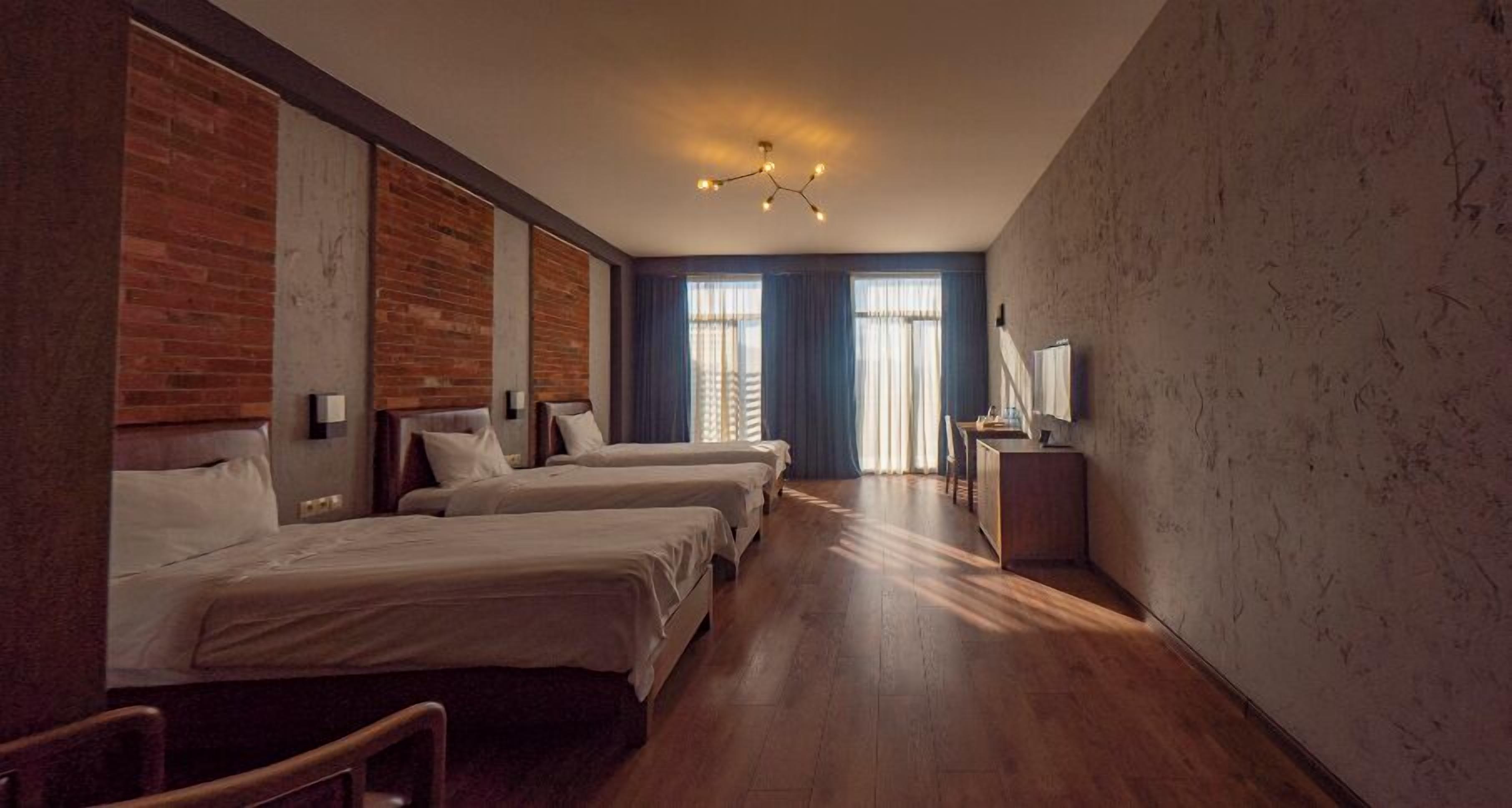 Une chambre d’hôtel avec trois lits, un plancher en bois, un bureau avec une chaise et une grande fenêtre avec des rideaux.