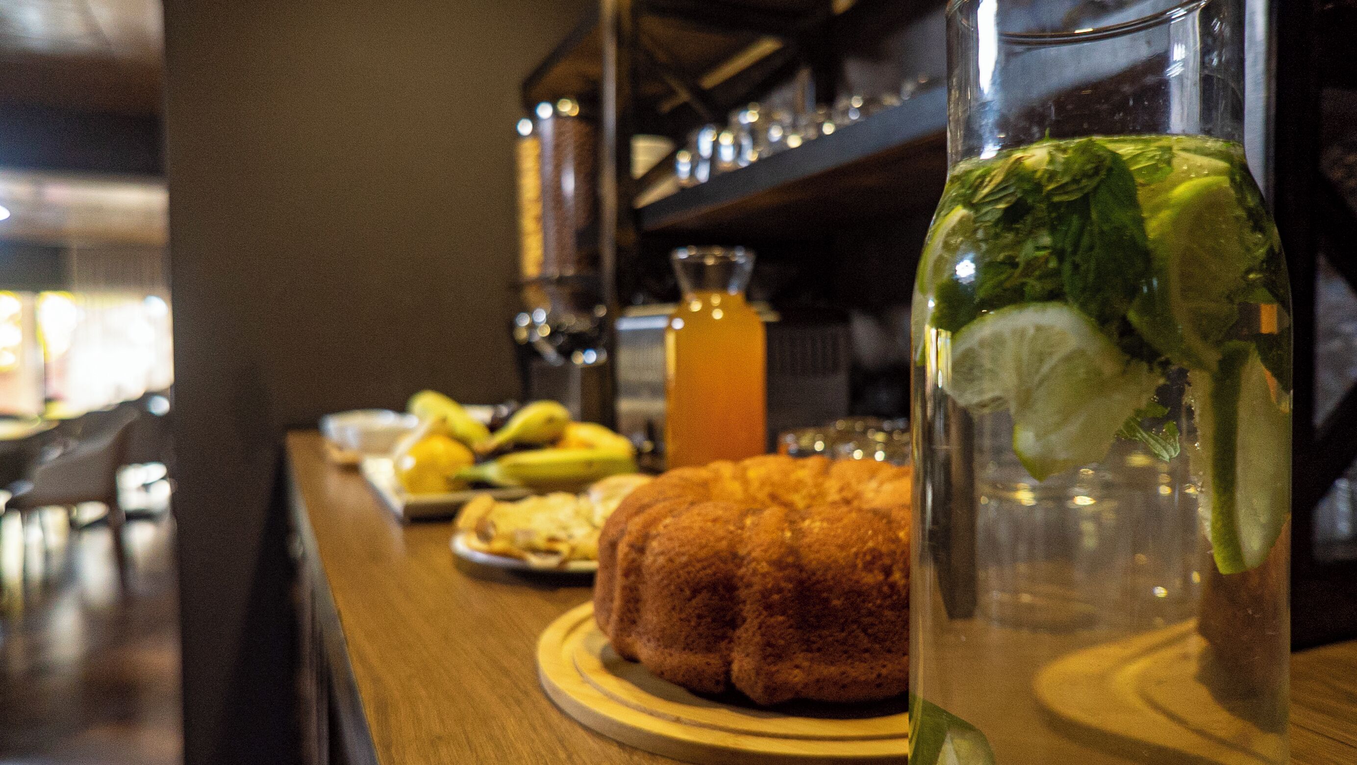 A bar setting with a glass of iced beverage, a plate of bananas, and a bundt cake on a wooden table.