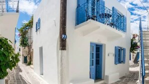 A white building with blue shutters and a balcony, a narrow street, and a sign indicating a parking area. - Au Cœur de Parikia , Appartement Cosy Avec Balcon (Parikiá)