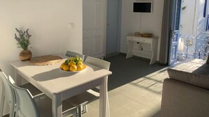 A bright, modern kitchen with white cabinetry, a dining table with a bowl of fruit, and a TV mounted above a white cabinet. - Au Cœur de Parikia , Appartement Cosy Avec Balcon (Parikiá)