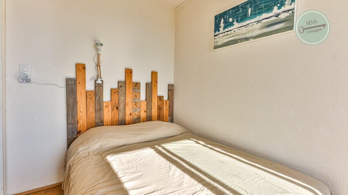 A bedroom with a wooden headboard, a bed with a beige cover, a wall-mounted light fixture, and a painting of a snowy landscape.