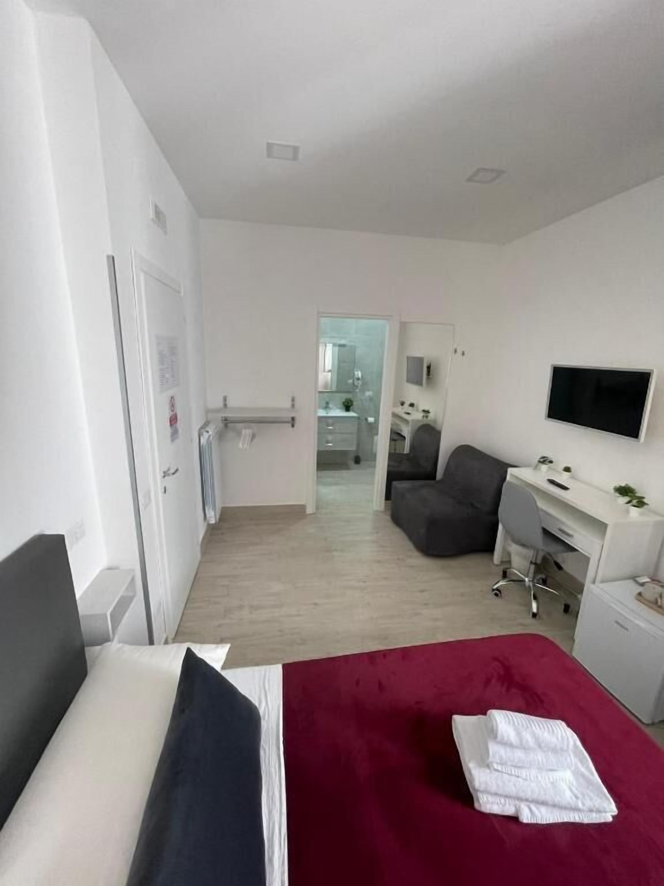 A hotel room with a bed, a desk, a chair, a TV, and a bathroom visible through an open door.