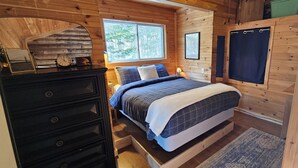 A cozy bedroom with a wooden interior, a bed with a blue and white checkered duvet, a dresser, a window with a view of trees, and a framed picture on the wall. - Overlook Cabin - Lake/Mountain View*Pet Friendly*A/C*Washer/Dryer (Greenville)