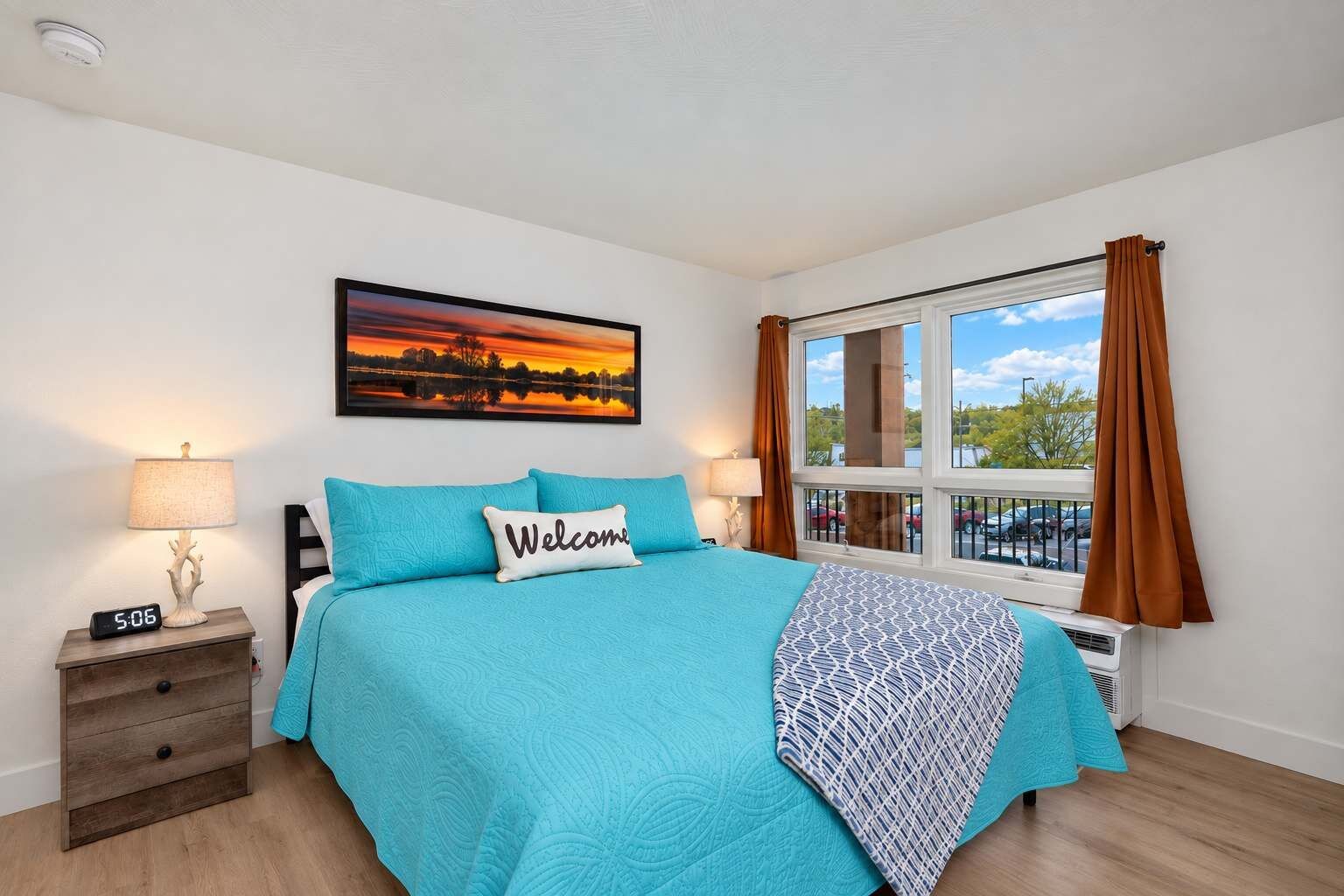 A bedroom with a bed, two bedside tables, a nightstand, a wall clock, a window with curtains, and a painting.