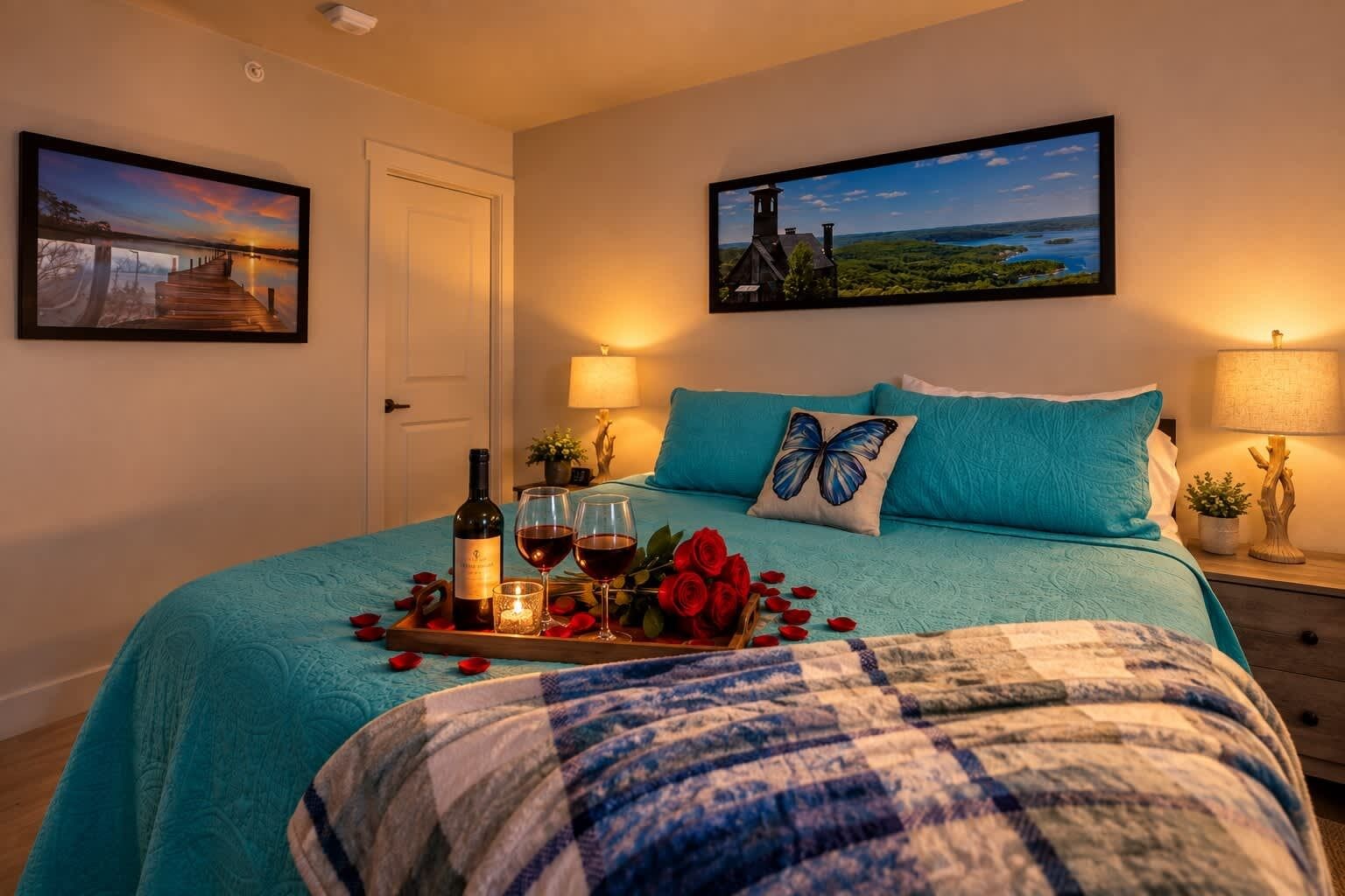 A bedroom with a bed, a nightstand, a lamp, a tray with wine, roses, and a candle.