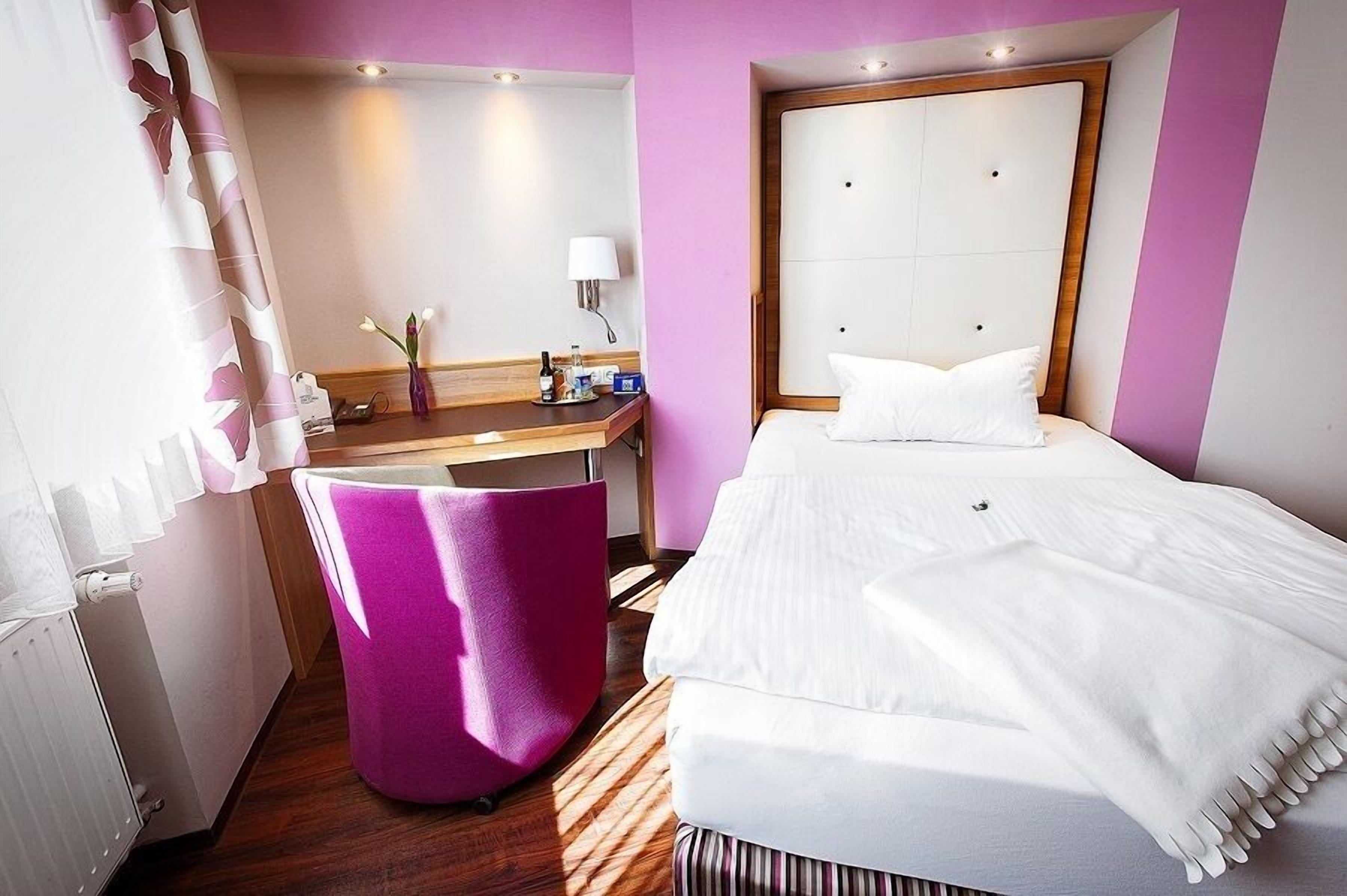 Une chambre d’hôtel avec un lit, un bureau, une chaise, un miroir et une fenêtre avec des rideaux.