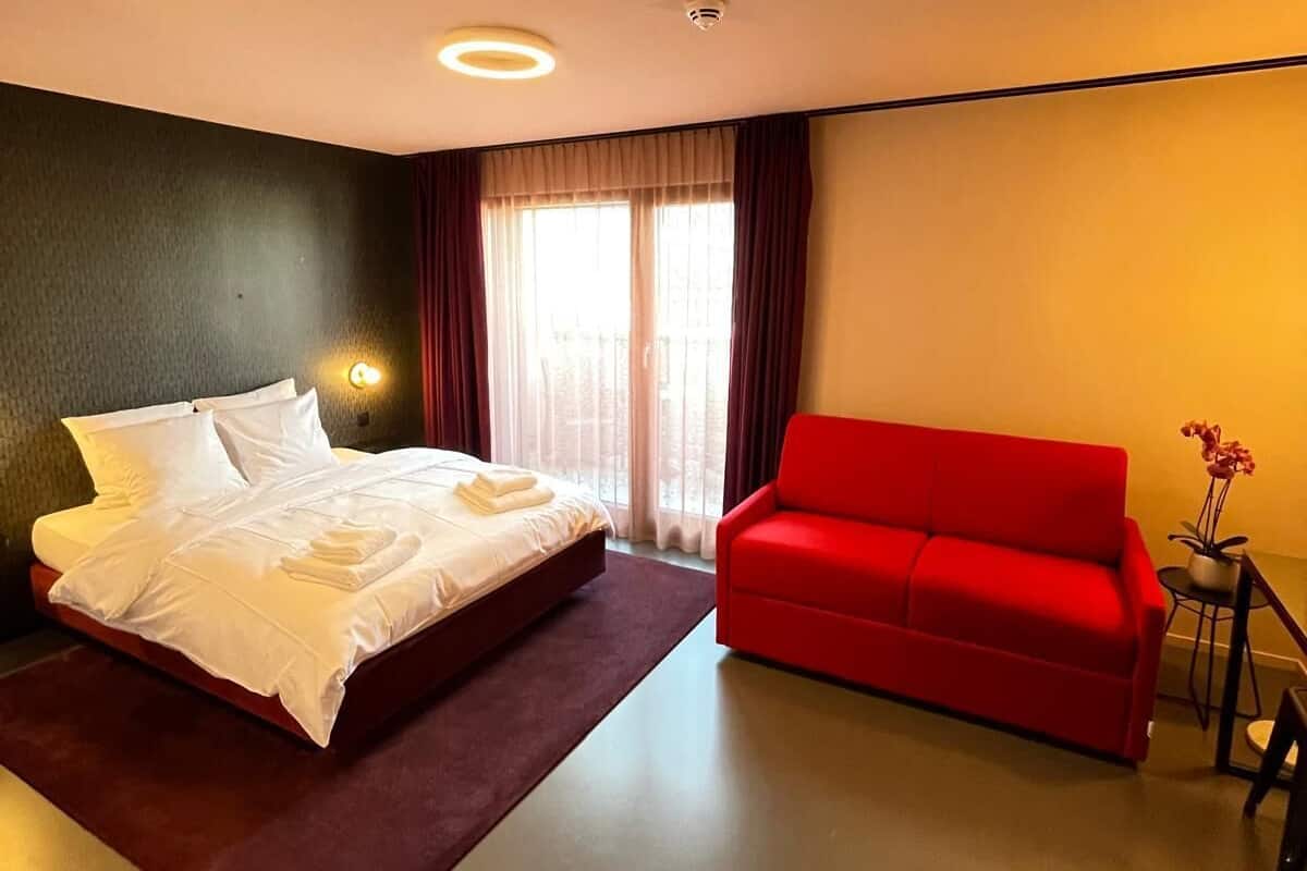 A hotel room with a bed, a red sofa, a small table with a plant, and a window with curtains.