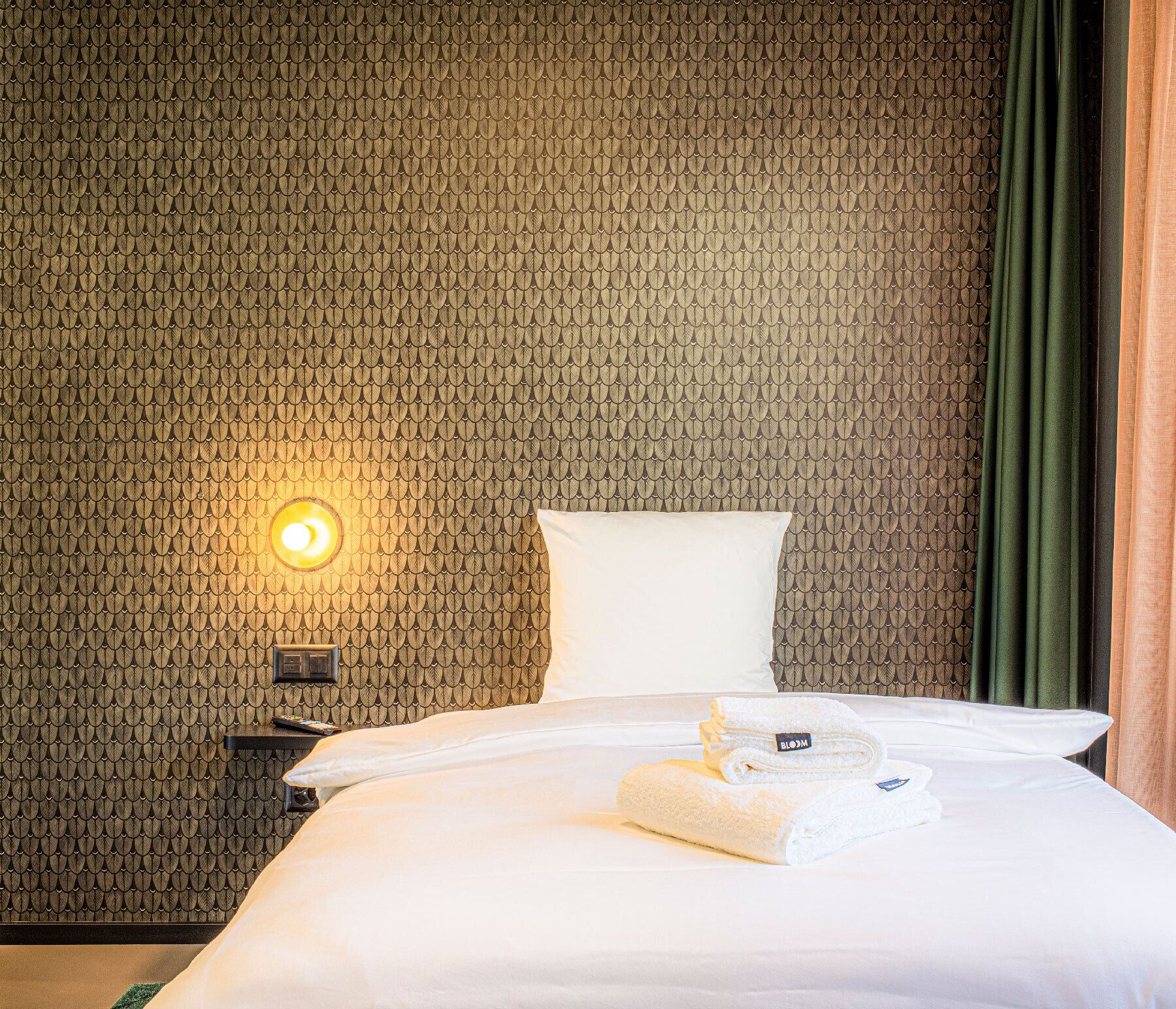 A hotel room with a patterned wall, a bed with white linens, a bedside table with a lamp, and a green curtain.