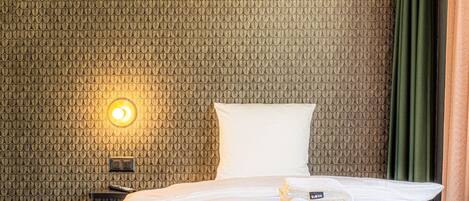A hotel room with a patterned wall, a bed with white linens, a bedside table with a lamp, and a green curtain.
