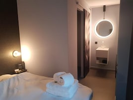 Una habitación de hotel con una cama bien tendida, un espejo redondo sobre el lavabo y una mesita de noche con una lámpara.