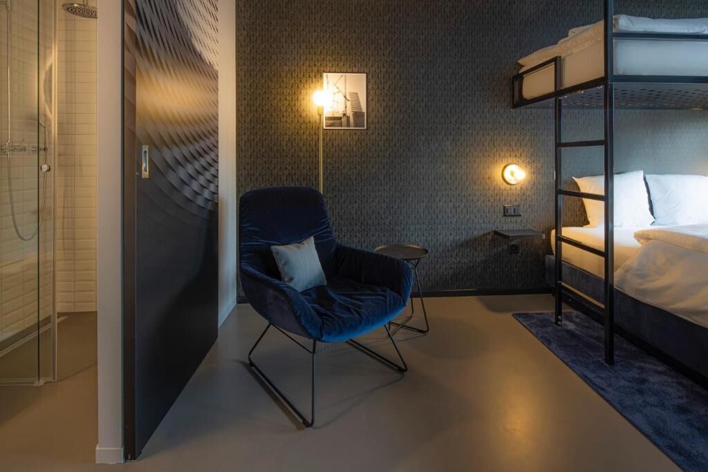 A modern hotel room with a bunk bed, a blue armchair, a small table, and a wall-mounted light.