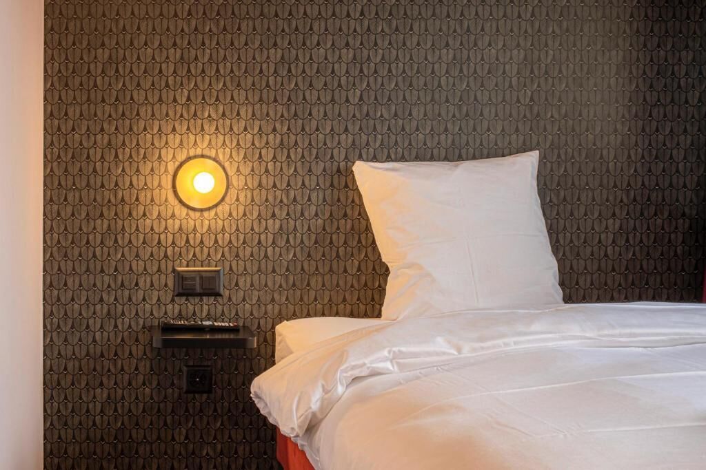 Habitación de hotel con pared estampada, cama con ropa blanca y lámpara de pared.