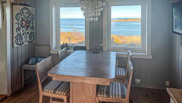 A dining area with a wooden table and chairs, two windows with a view of the ocean, and a chandelier. - Nice home in Smøla with house sea view (Smøla)