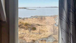 A view through a window of a coastal landscape with dry grass, rocky terrain, and a calm sea. - Nice home in Smøla with house sea view (Smøla)