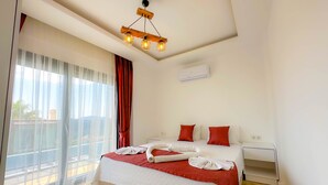 Quarto de hotel com cama, mesa de cabeceira, janela com cortinas leves e luminária no teto.