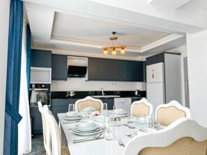 A dining area with a white table set for four, surrounded by chairs with a beige and white pattern. A modern chandelier hangs above the table. - Nokta Park J (Fethiye)