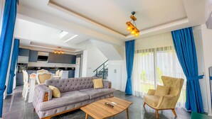 Uma sala de estar ampla com sofá, poltrona e mesa de centro, uma cozinha ao fundo e grandes janelas com cortinas.