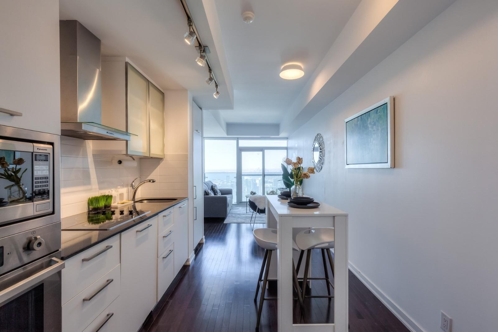 A modern kitchen with a built-in oven, a dining area with a white table and bar stools, and a view of the city through large windows.