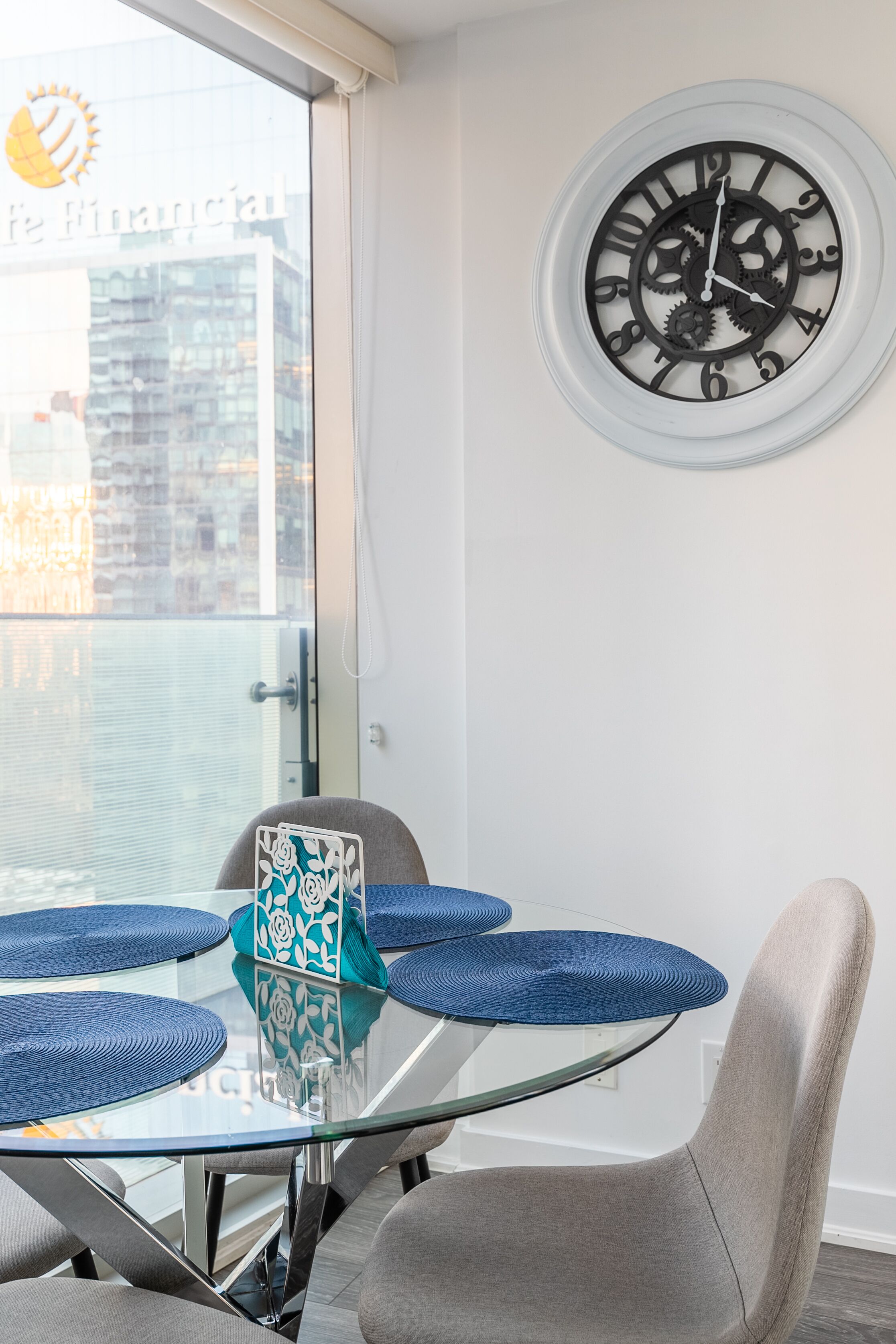 A modern dining area with a glass table, grey chairs, and a large wall clock.