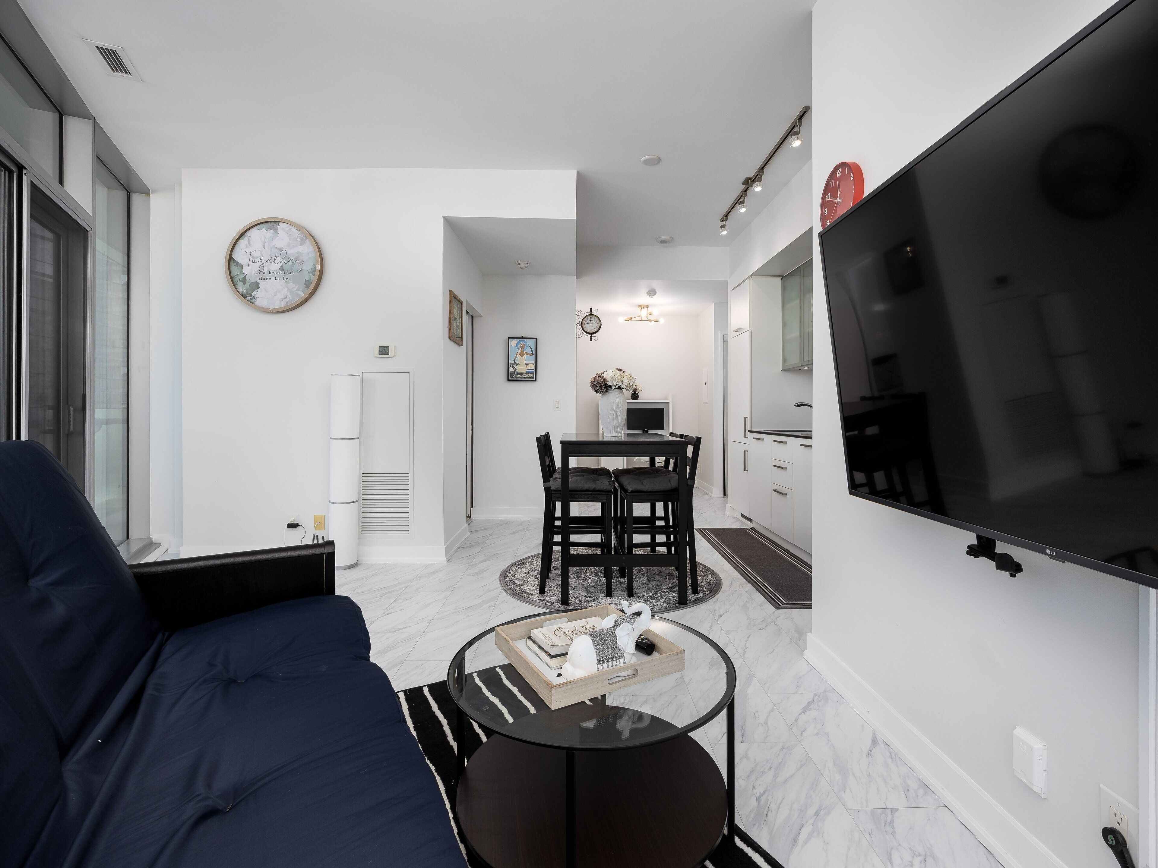 Una sala de estar moderna con un sofá, una mesa de centro y un televisor. Hay una zona de comedor con una mesa y sillas. La habitación tiene paredes blancas y un ventanal grande.
