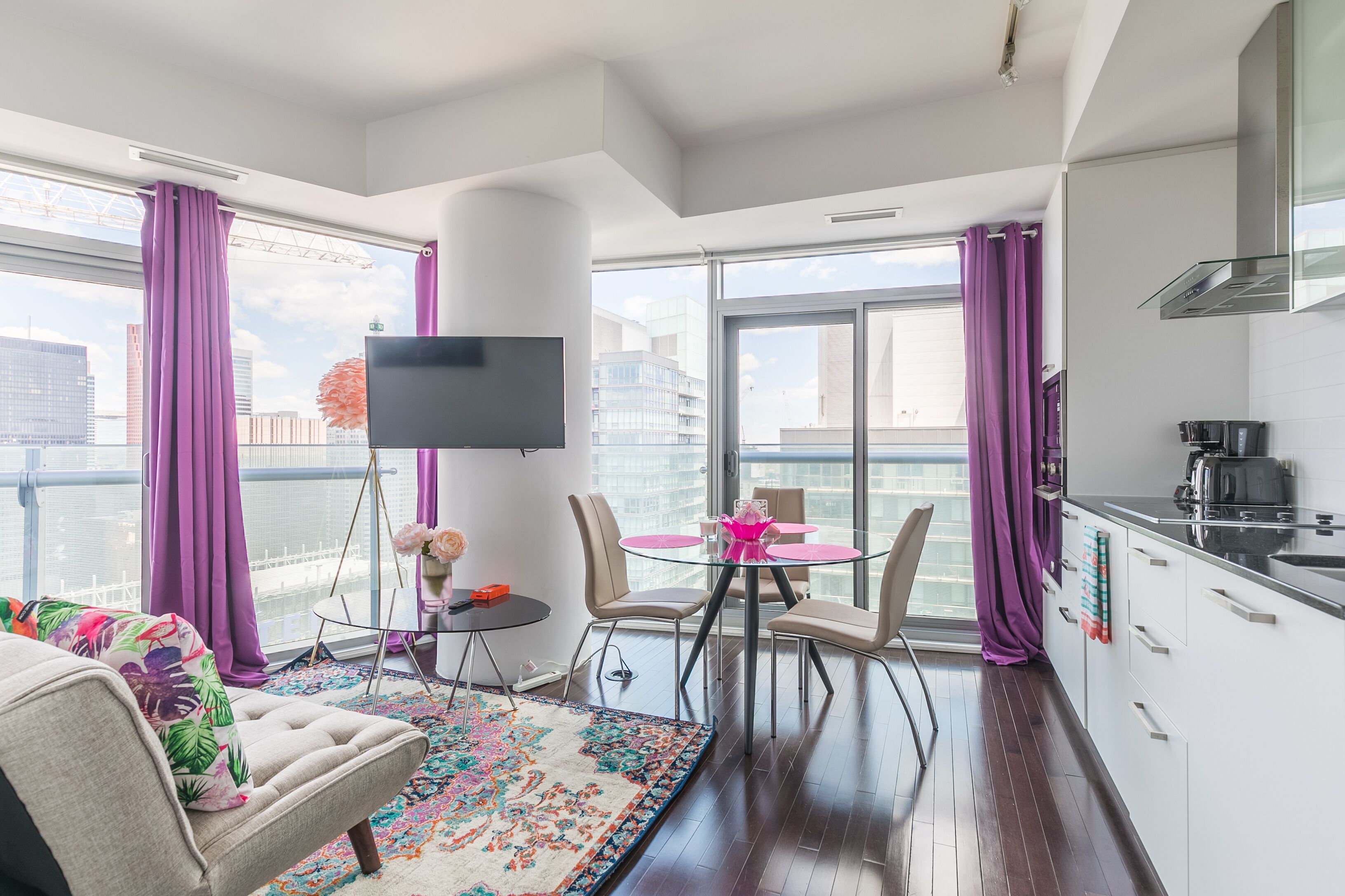 Een moderne woonkamer met een bank, een salontafel en een televisie. Er is een balkon met paarse gordijnen en uitzicht op de stad.