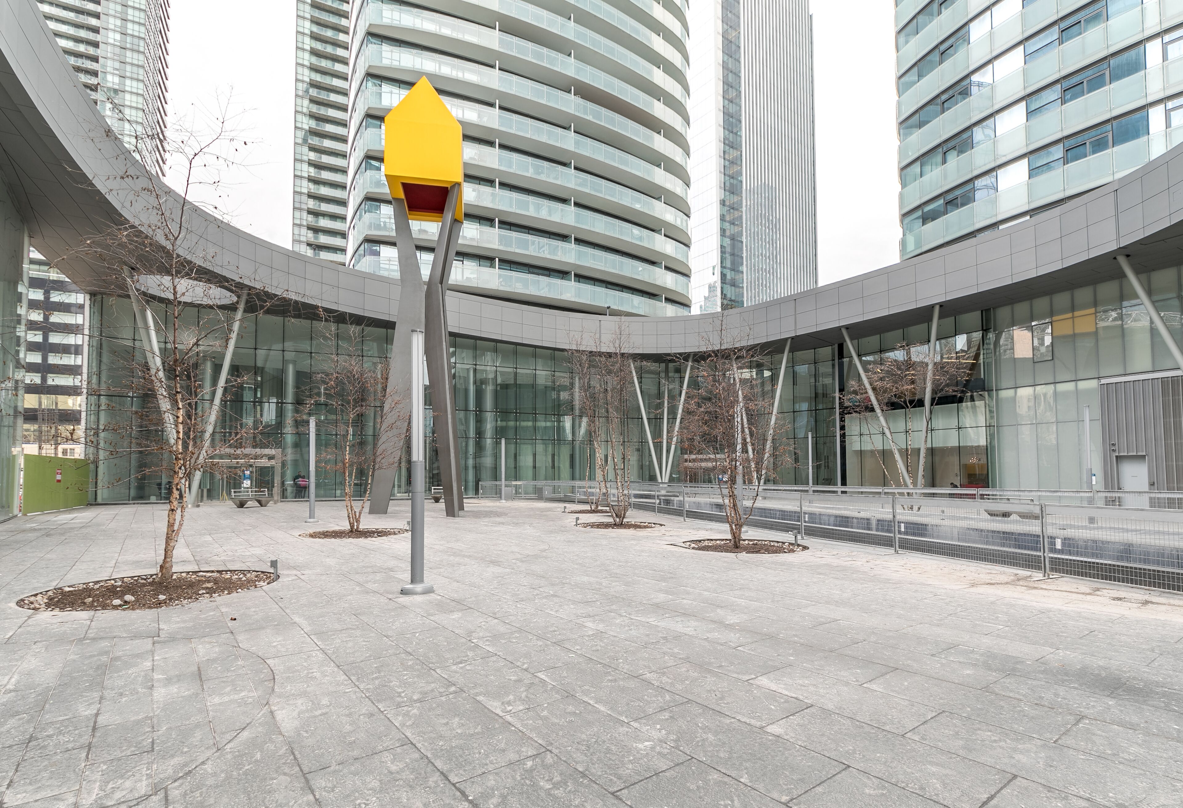 Une place urbaine moderne, avec un bâtiment aux façades courbes en verre, un panneau de signalisation jaune et des arbres sans feuilles.