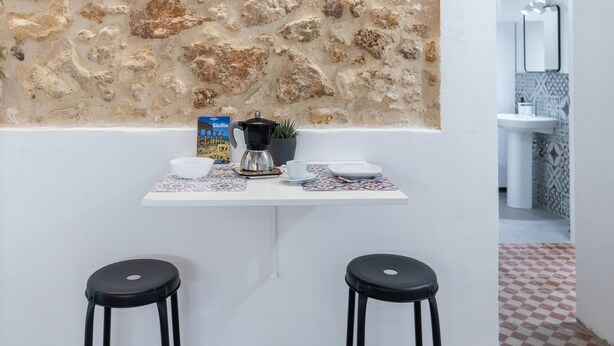 A kitchen with a stone wall, a coffee maker, a bowl, a cup, and two stools. - Dimora Netina by Wonderful Italy (Noto)
