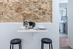 Una cocina con pared de piedra, una cafetera, un bol, una taza y dos taburetes.