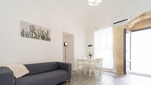 A living room with a gray sofa, a dining table with chairs, a cactus artwork on the wall, and a large window with a view. - Dimora Netina by Wonderful Italy (Noto)